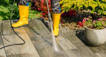 A person wearing yellow rubber boots uses a pressure washer on a stone patio.