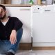 Man sitting on the floor leaning back on apliances and holding his forhead.